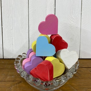 May include: A heart-shaped glass dish filled with colorful wooden hearts. The hearts are painted in shades of pink, red, blue, yellow, and white. The dish is sitting on a wooden surface.