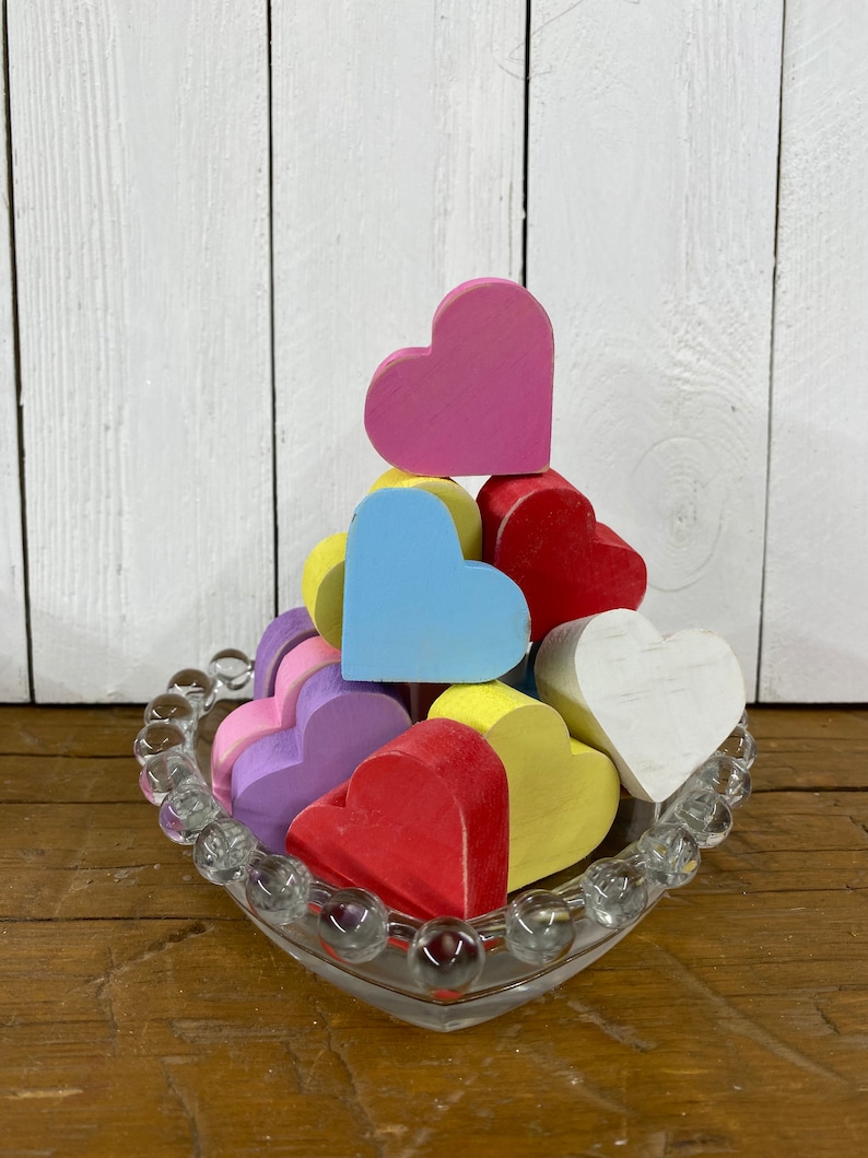 May include: A heart-shaped glass dish filled with colorful wooden hearts. The hearts are painted in shades of pink, red, blue, yellow, and white. The dish is sitting on a wooden surface.