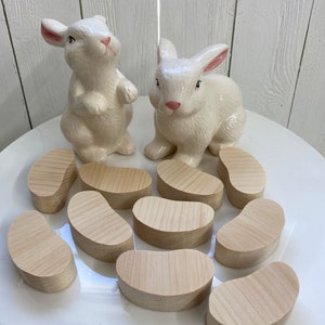 May include: Ten wooden, light brown, bean-shaped, unfinished, wooden pieces on a white plate with two white ceramic bunny figurines.