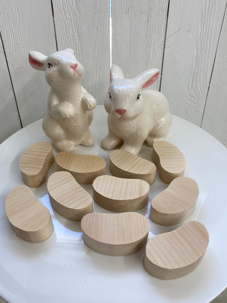 May include: Ten wooden, light brown, bean-shaped, unfinished, wooden pieces on a white plate with two white ceramic bunny figurines.