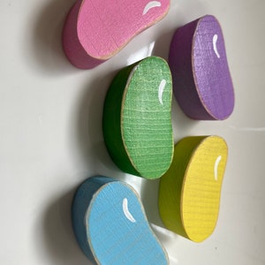 May include: Five wooden jelly bean shaped blocks in different colors: pink, purple, green, yellow, and blue. Each block has a white painted line on it.