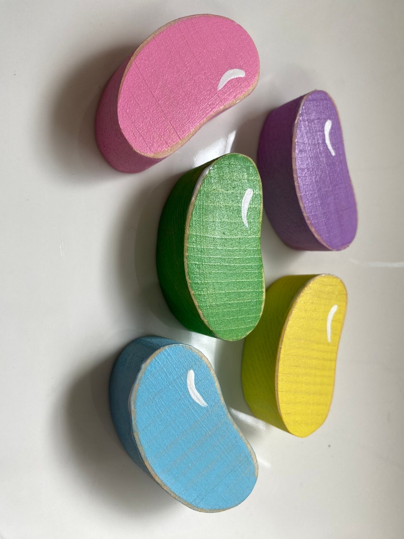 May include: Five wooden jelly bean shaped blocks in different colors: pink, purple, green, yellow, and blue. Each block has a white painted line on it.