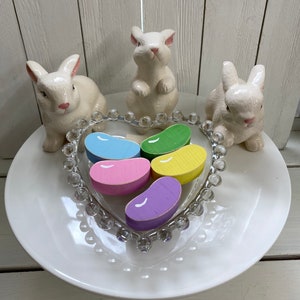 May include: Five wooden jelly bean shaped decorations in a heart shaped glass dish. The jelly beans are in shades of pink, purple, yellow, green, and blue. Three white ceramic bunny figurines surround the dish on a white plate.