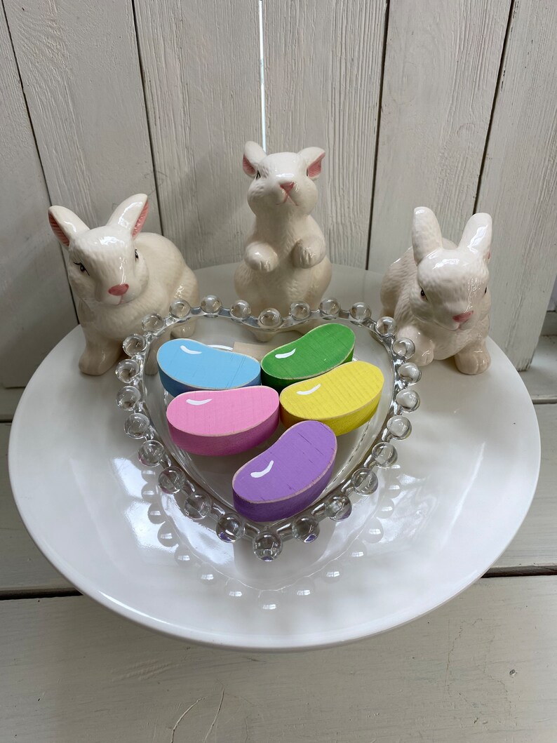 May include: Five wooden jelly bean shaped decorations in a heart shaped glass dish. The jelly beans are in shades of pink, purple, yellow, green, and blue. Three white ceramic bunny figurines surround the dish on a white plate.