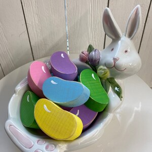 May include: A white ceramic bunny figurine holding a bowl filled with colorful wooden jelly bean shaped candies.