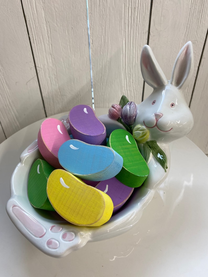 May include: A white ceramic bunny figurine holding a bowl filled with colorful wooden jelly bean shaped candies.