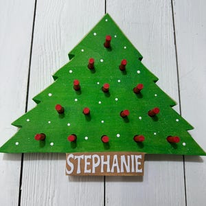 May include: A green, Christmas tree-shaped wooden decoration with red pegs and white polka dots. The name "STEPHANIE" is displayed on a small wooden sign at the base. The tree is mounted on a white wooden surface.