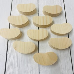 May include: Ten unfinished wooden shapes in the form of jelly beans. The shapes are all the same size and are arranged in a random pattern on a white wooden surface.