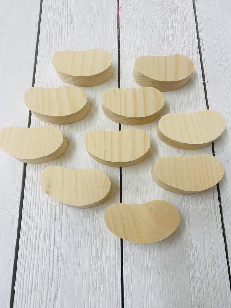 May include: Ten unfinished wooden shapes in the form of jelly beans. The shapes are all the same size and are arranged in a random pattern on a white wooden surface.