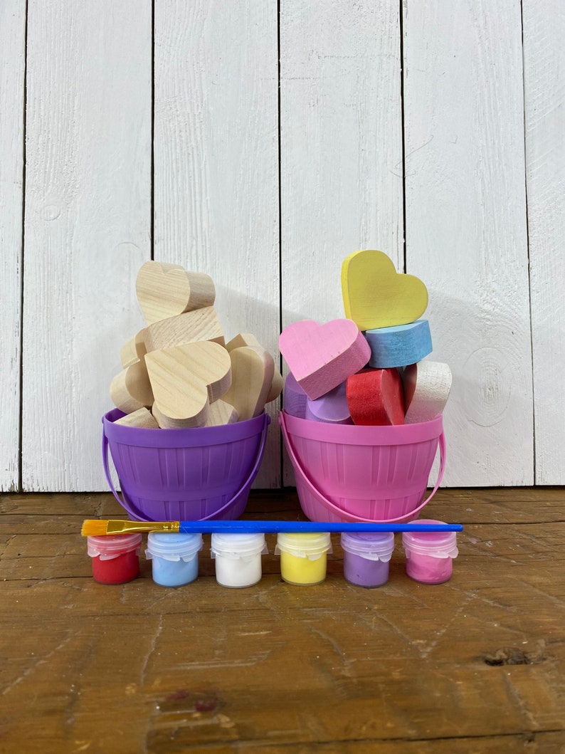 May include: Two buckets filled with wooden heart shapes, one purple bucket with natural wood hearts and one pink bucket with painted hearts in pink, yellow, blue and red.  A paintbrush and six small paint bottles in red, blue, white, yellow, purple and pink are in front of the buckets.