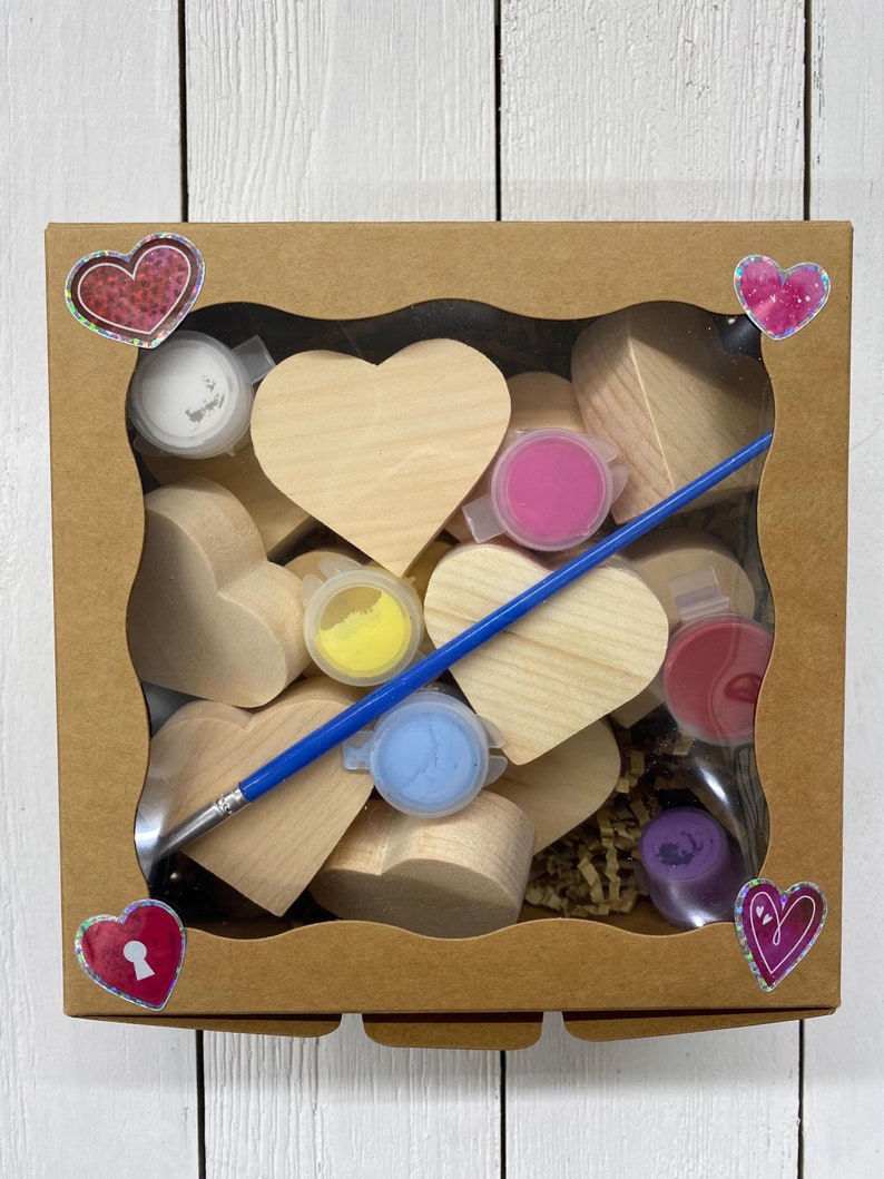 May include: A craft kit with wooden heart shapes, paint, and a paintbrush in a brown cardboard box. The box has heart-shaped stickers with glitter.