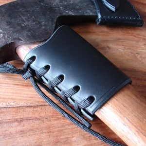 May include: A black leather axe sheath with a lace-up closure. The sheath is wrapped around the wooden handle of an axe.