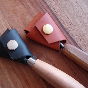 Puede incluir: Dos portaherramientas de cuero con broches metálicos. Los portaherramientas son marrones y negros y están envueltos alrededor de los mangos de dos herramientas de talla de madera.