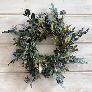 May include: A green and blue wreath with small purple flowers. The wreath is made of artificial greenery and is perfect for a spring or summer decor.