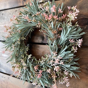 May include: A green and pink artificial wreath with small pink flowers and green leaves. The wreath is hanging on a wooden wall.