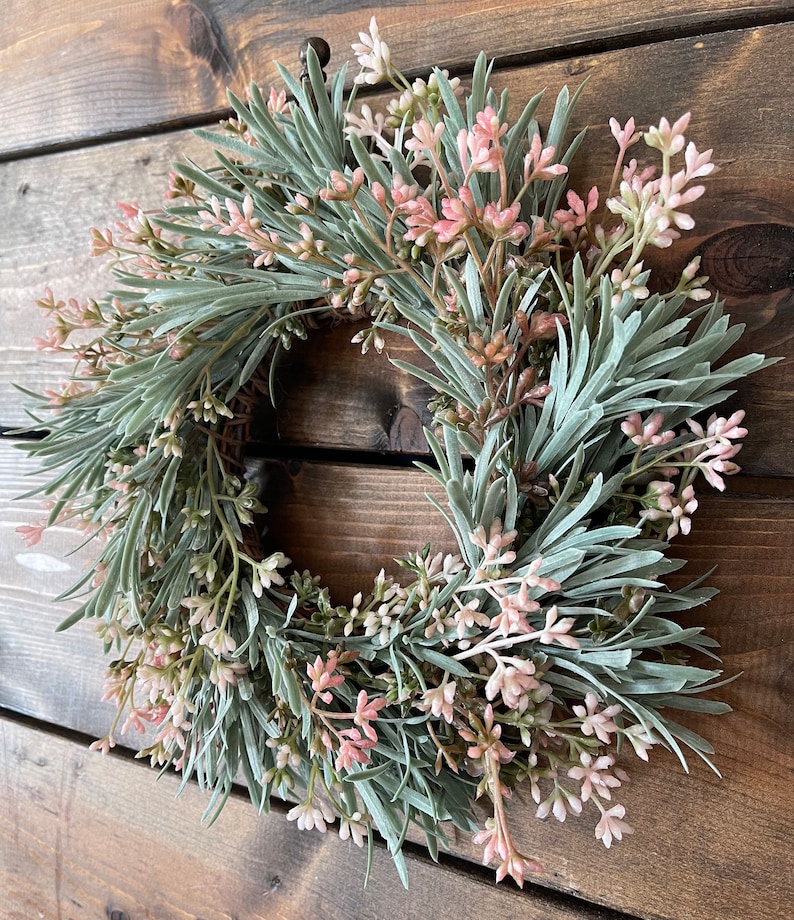 May include: A green and pink artificial wreath with small pink flowers and green leaves. The wreath is hanging on a wooden wall.
