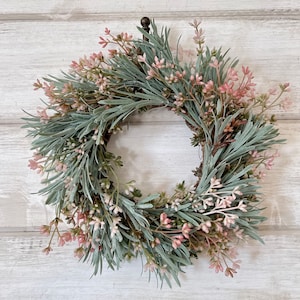 May include: A green and pink artificial wreath with a metal hook. The wreath is made of sprigs of greenery with small pink flowers.