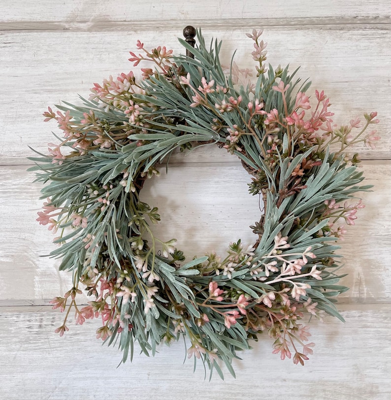 May include: A green and pink artificial wreath with a metal hook. The wreath is made of sprigs of greenery with small pink flowers.