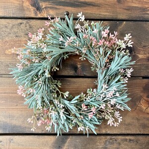 May include: A green and pink artificial wreath with small sprigs of greenery and pink flowers. The wreath is hanging on a wooden surface.