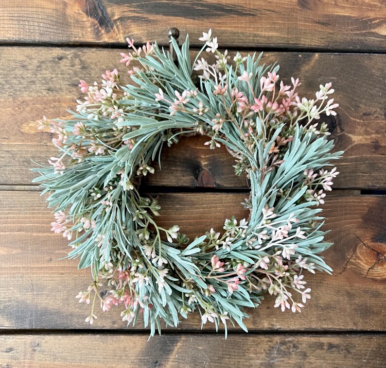 May include: A green and pink artificial wreath with small sprigs of greenery and pink flowers. The wreath is hanging on a wooden surface.