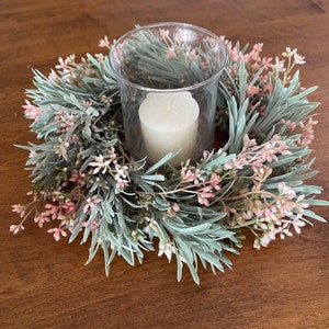 May include: A white candle in a clear glass holder surrounded by a green and pink floral wreath.