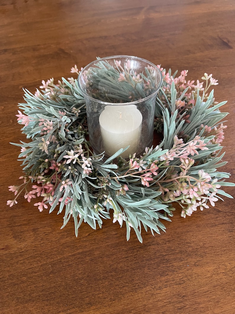 May include: A white candle in a clear glass holder surrounded by a green and pink floral wreath.