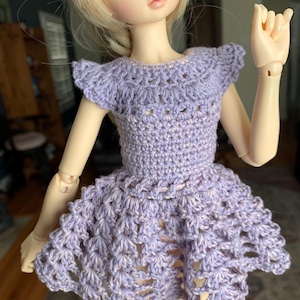 May include: A lavender crochet dress with a flared skirt and cap sleeves. The doll-sized dress is handmade with a delicate openwork pattern. The dress is designed to fit a doll with articulated joints and a blonde wig.