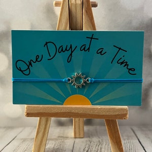May include: A blue elastic bracelet with a silver charm featuring a sun with a turquoise center. The bracelet is displayed on a small wooden easel with a card that reads "One Day at a Time".