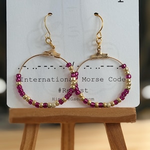 May include: Gold-tone hoop earrings adorned with magenta and gold beads, arranged in a Morse code pattern. The earrings hang from a card with the text "Fuck Trump" and "International Morse Code".