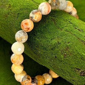 May include: A beaded bracelet featuring round, polished stones in shades of cream, orange, and gray. The bracelet is displayed on a textured green surface, showcasing the natural variations in the stone's patterns and colors. A piece of jewelry.