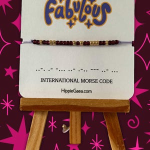 May include: A beaded bracelet with burgundy and gold beads is displayed on a small easel. The card behind the bracelet has the word "Fabulous" in blue script and Morse code. The website HippieGaea.com is also visible.