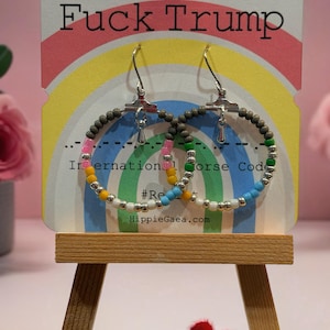 May include: A pair of hoop earrings featuring multi-colored beads in shades of grey, pink, green, yellow, blue, and white, accented with silver-toned metal. The earrings are displayed on a card with a rainbow graphic and the text Fuck Trump.
