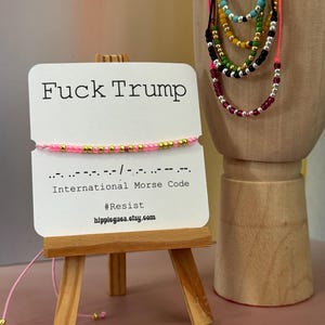 May include: A pink and gold beaded bracelet displayed on a small wooden easel. The card on the easel reads "Fuck Trump" and includes Morse code, the word "Resist," and a website address. Several colorful beaded necklaces are also visible.