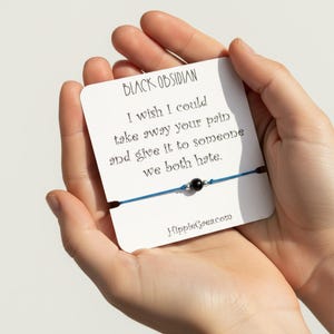 May include: A blue string bracelet with a single black obsidian bead and two small silver-toned metal beads, mounted on a white display card. The card features the text: BLACK OBSIDIAN, I wish I could take away your pain and give it to someone we both hate, and HippieGaea.com.