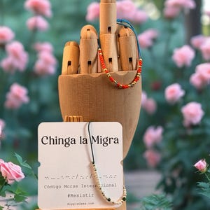 May include: A wooden hand model with two beaded bracelets. One bracelet has red and gold beads, the other has white, black, and gold beads. A small sign reads "Chinga la Migra" and "#Resistir".