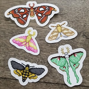 May include: A collection of five pixel art moth stickers. The stickers feature various moth designs in shades of red, yellow, pink, green, and black. Each sticker has a white border and is placed on a wooden surface.