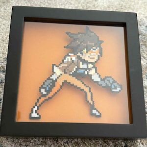 May include: Framed pixel art of a character from the video game Overwatch. The artwork features a figure in a dynamic pose, rendered in a pixelated style with orange, brown, white, and blue tones. The art is set against an orange background within a black frame.