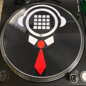 May include: A black turntable with a white and red DJ slipmat featuring a stylized illustration of a person wearing headphones and a tie. The slipmat has a grid pattern on the head and a red tie.