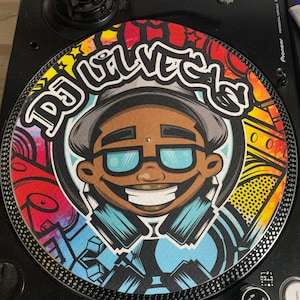 Pode incluir: Um slipmat colorido para toca-discos com um personagem de DJ de desenho animado e o texto "DJ Lil'Vets". O design incorpora cores vibrantes e elementos de estilo graffiti, em um toca-discos preto.