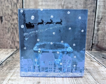Op de afbeelding: Een blauwe vierkante kandelaar met een winters tafereel. Het ontwerp toont een silhouet van de slee en rendieren van de kerstman, sneeuwvlokken en een dorp. De kandelaar is gemaakt van glas en heeft een glitterende basis.