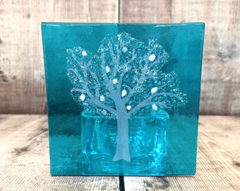 Op de afbeelding: Een vierkante, turquoise glazen kandelaar met een mat boomontwerp. De boom heeft witte accenten en is gecentreerd op de voorkant. De kandelaar is ongeveer 10 cm hoog en 10 cm breed.