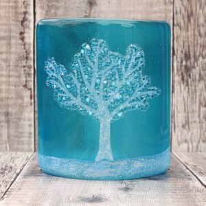 May include: A teal glass candle holder featuring a white winter tree design with sparkling details. The teal background complements the frosted base, creating a snowy effect. The candle holder is cylindrical in shape.