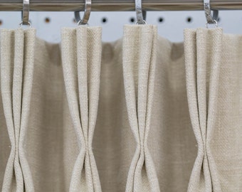 May include: Close-up of four beige curtains with pleats hanging from a silver curtain rod.
