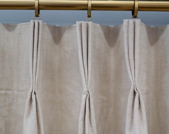 May include: Close-up of a set of three beige linen curtains with pleats hanging from a gold metal curtain rod.