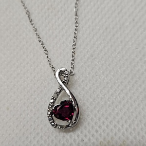 May include: A silver-toned necklace featuring an infinity-shaped pendant. The pendant is accented with small clear stones along one side and holds a deep red, heart-shaped gemstone in the center. The chain is a delicate, thin silver-toned link style.