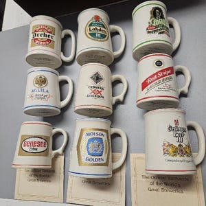 May include: A collection of eight white ceramic beer mugs, each featuring a unique brewery logo and name, including Dreher, Lahden, Cerveza Dos Equis, Red Stripe, Aguila, Genesee, Molson Golden, and Oranjeboom, with some mugs resting on paper labels.