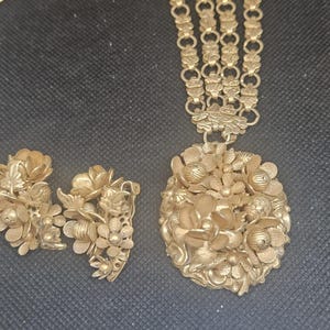 May include: A gold-toned jewelry set consisting of a necklace and a pair of clip-on earrings. The necklace features a multi-strand chain leading to a large, round pendant. Both the pendant and the earrings are decorated with detailed floral motifs.