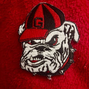 May include: An embroidered patch featuring a white bulldog head with a black and red spiked collar, wearing a black and red baseball cap with a prominent letter G on the front. The design uses black, white, and red thread on a textured fabric.