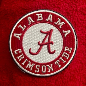 Könnte beinhalten: Ein kreisförmiger gestickter Aufnäher mit weißer Mitte und einem roten geschriebenen Buchstaben A. Der umlaufende rote Rand enthält den weißen Text ALABAMA CRIMSON TIDE. Der Aufnäher ist auf einem strukturierten roten Stoff mit flauschiger Optik befestigt.