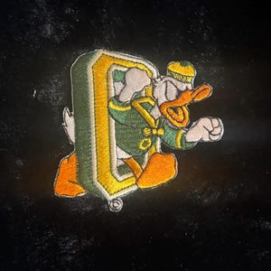 May include: An embroidered patch featuring a cartoon duck wearing a green and yellow jacket and a matching cap, stepping through a large, three-dimensional yellow and green letter O. The duck has orange feet and a beak, with white feathers.